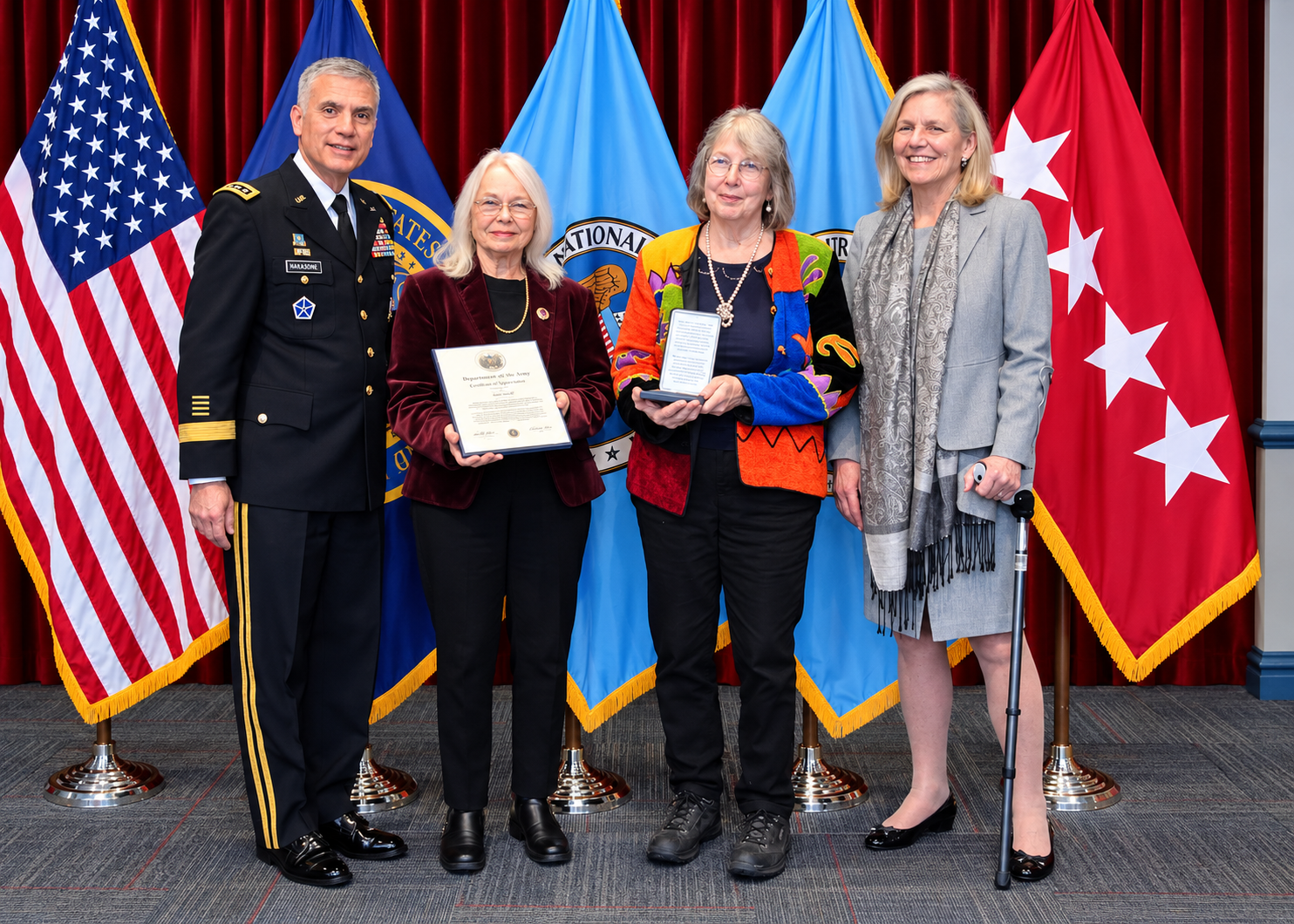 Gen. Paul M. Nakasone in Army dress uniform stands with Jean C. O'Connor, who holds a framed certificate, and two other women, one holding a medal, in front of the American flag and NSA flags